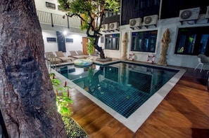 A swimming pool with a tree and lounge chairs, surrounded by a wooden deck and a white wall with air conditioning units. - Sathu Hotel (Chiang Mai)