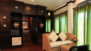 A room with dark wood paneling, a white refrigerator, a wicker sofa with cushions, a glass coffee table, and green curtains. - Sathu Hotel (Chiang Mai)
