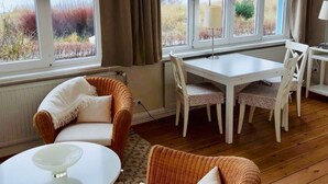 A room with a dining table and chairs, a wicker sofa, and a view of the outdoors through large windows. - Kleines Strandhotel (Timmendorfer Strand)
