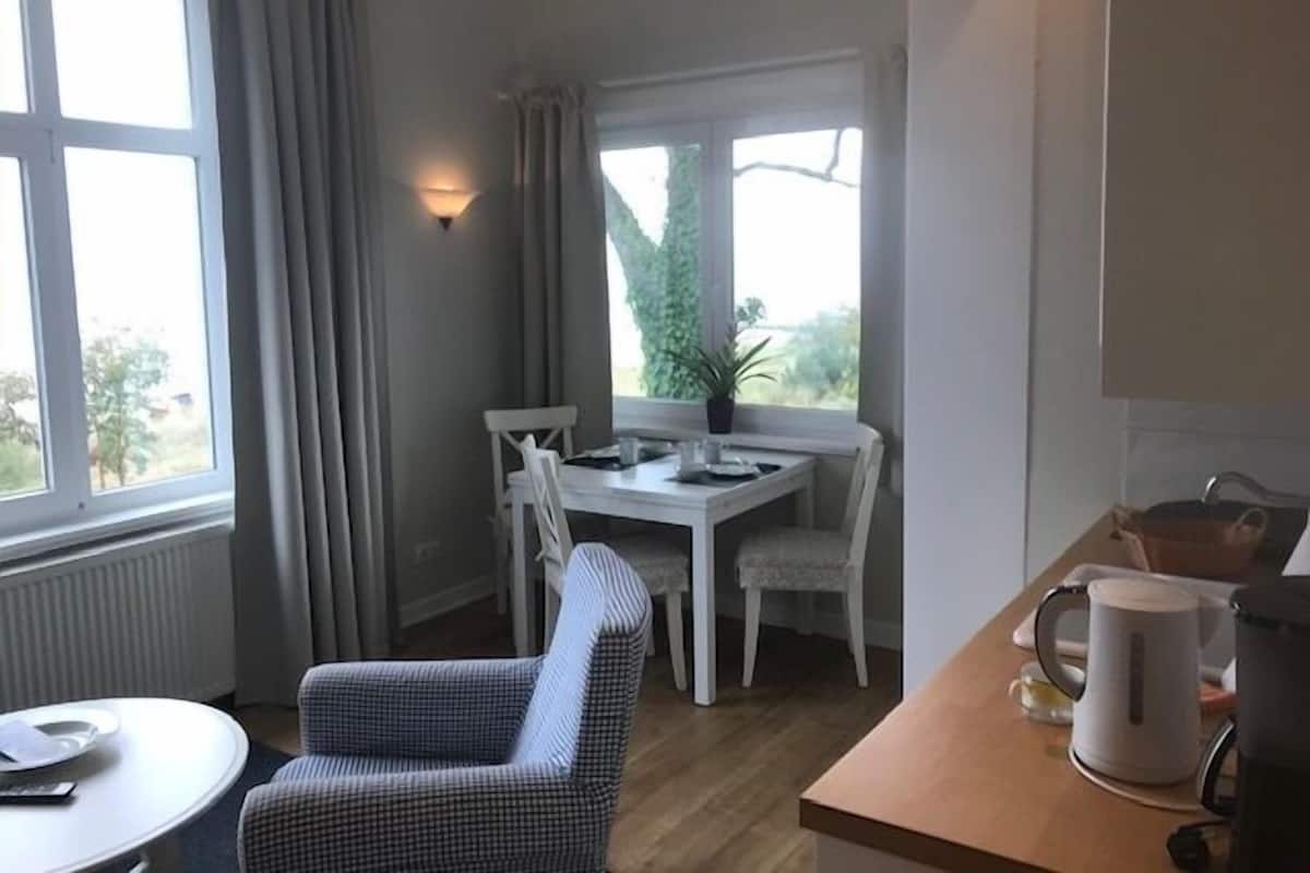 A small, well-lit room with a dining table and chairs, a single armchair, a radiator, and a window with curtains.