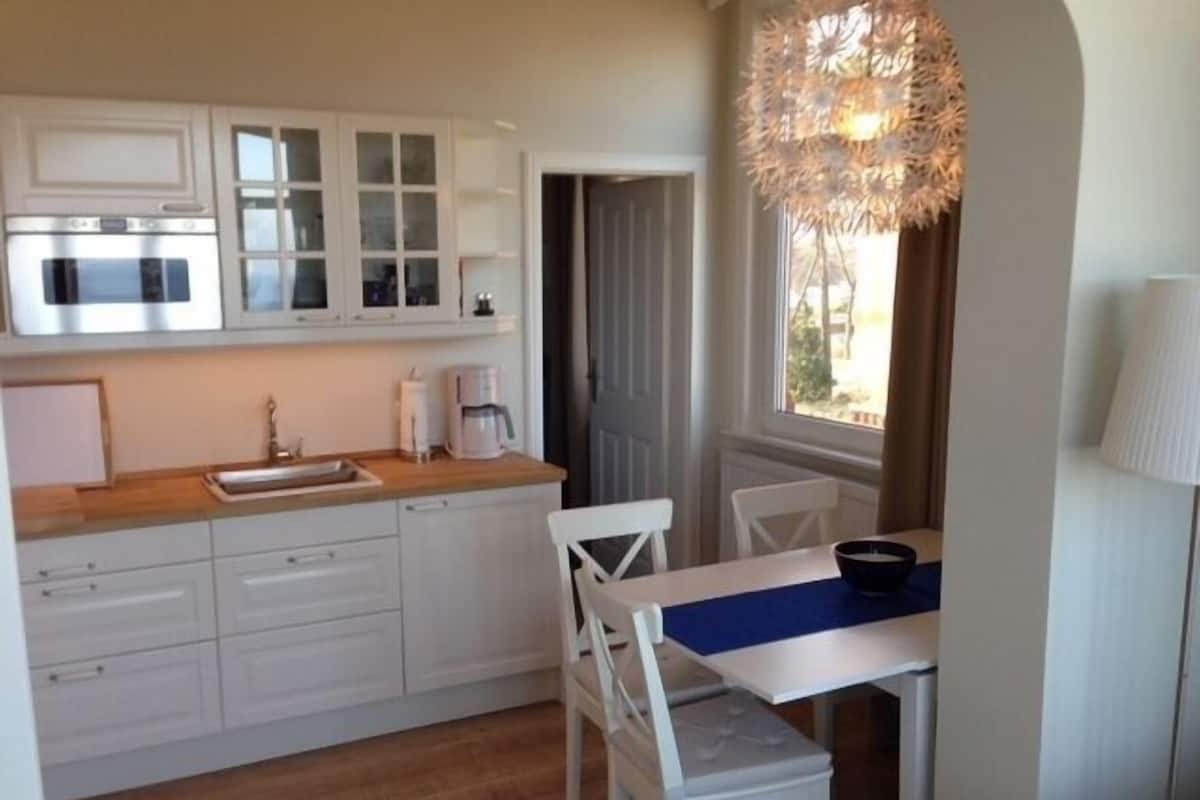 A compact kitchen with white cabinets, a microwave, and a coffee maker. There is a dining table with chairs and a lamp.