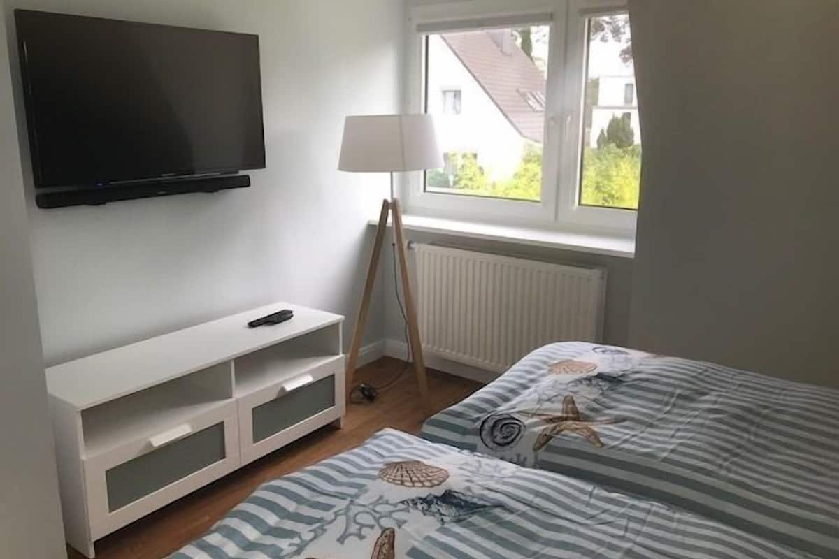 A bedroom with a TV mounted on the wall, a bedside table, a lamp, and a window with a view of the outside.