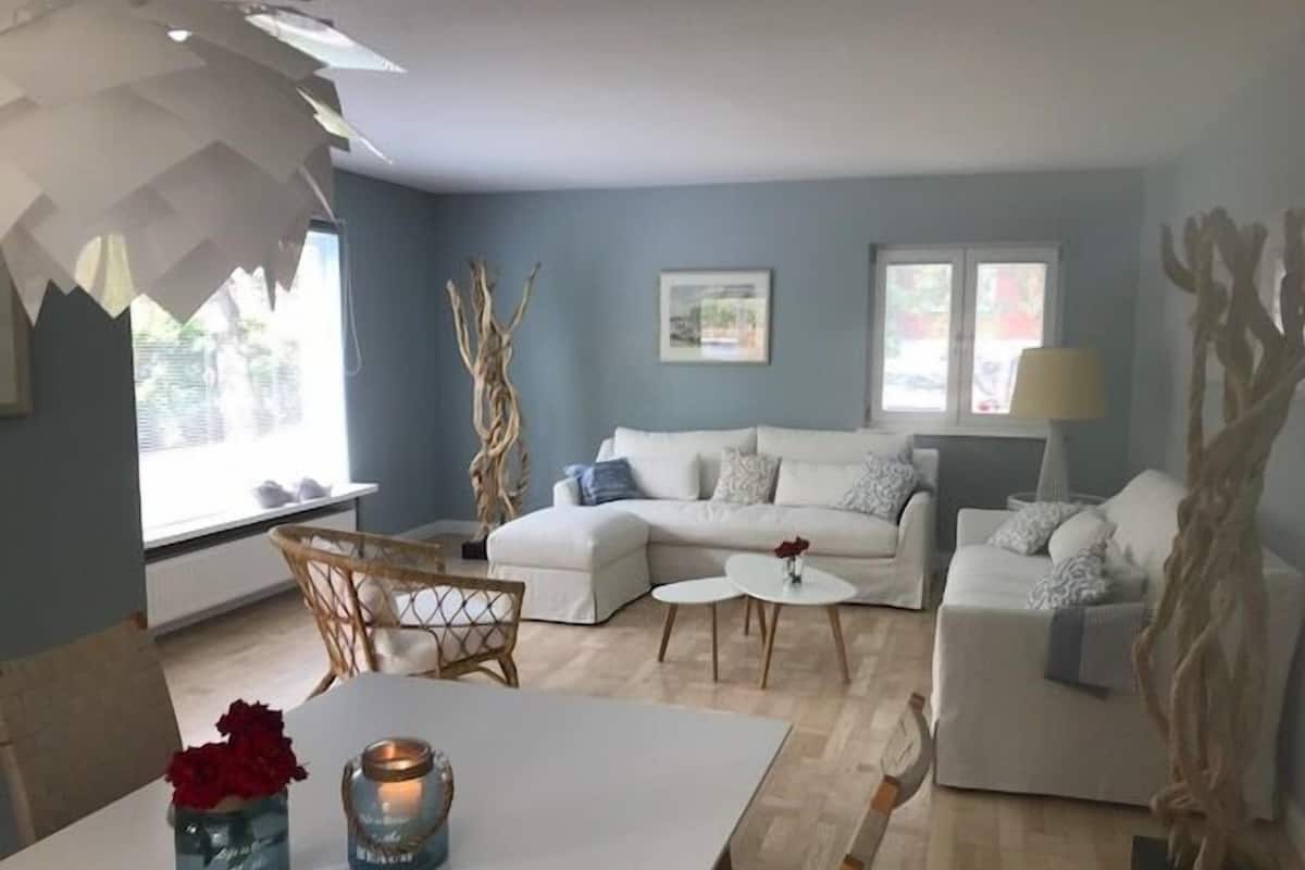 A modern living room with a white sofa, a small white table, and a dining area with a white table and chairs. There are decorative branches and a lamp on the side.