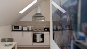 A modern kitchen with a microwave, toaster, kettle, and oven. There is a dining table with a glass of wine. The kitchen has a sloped ceiling with skylights. - Kirkstall House (Leeds)