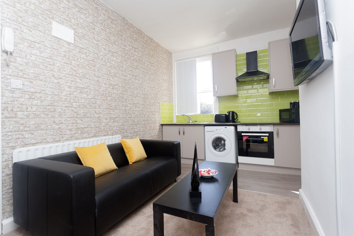 A compact living space with a black sofa, a coffee table, a washing machine, and a kitchen area with a microwave and a stove.