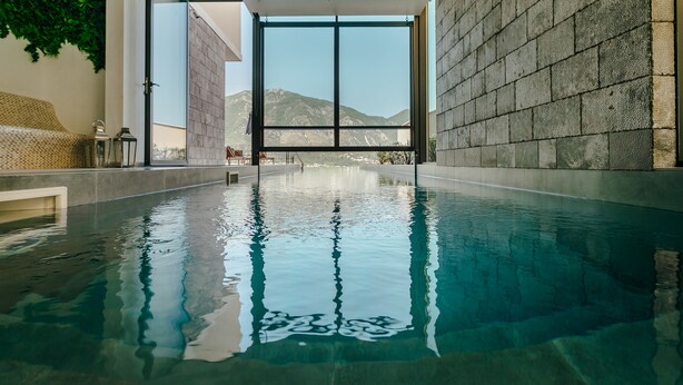 Une piscine intérieure à l’eau claire, avec vue sur les montagnes grâce à de grandes fenêtres, et un mur de pierre sur l’un des côtés.