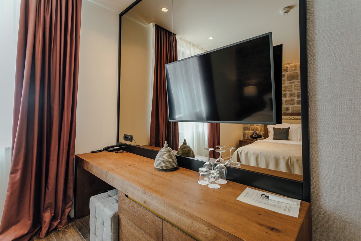 A hotel room with a wooden desk, a flat-screen TV, a bed with white linens, and a brick wall.