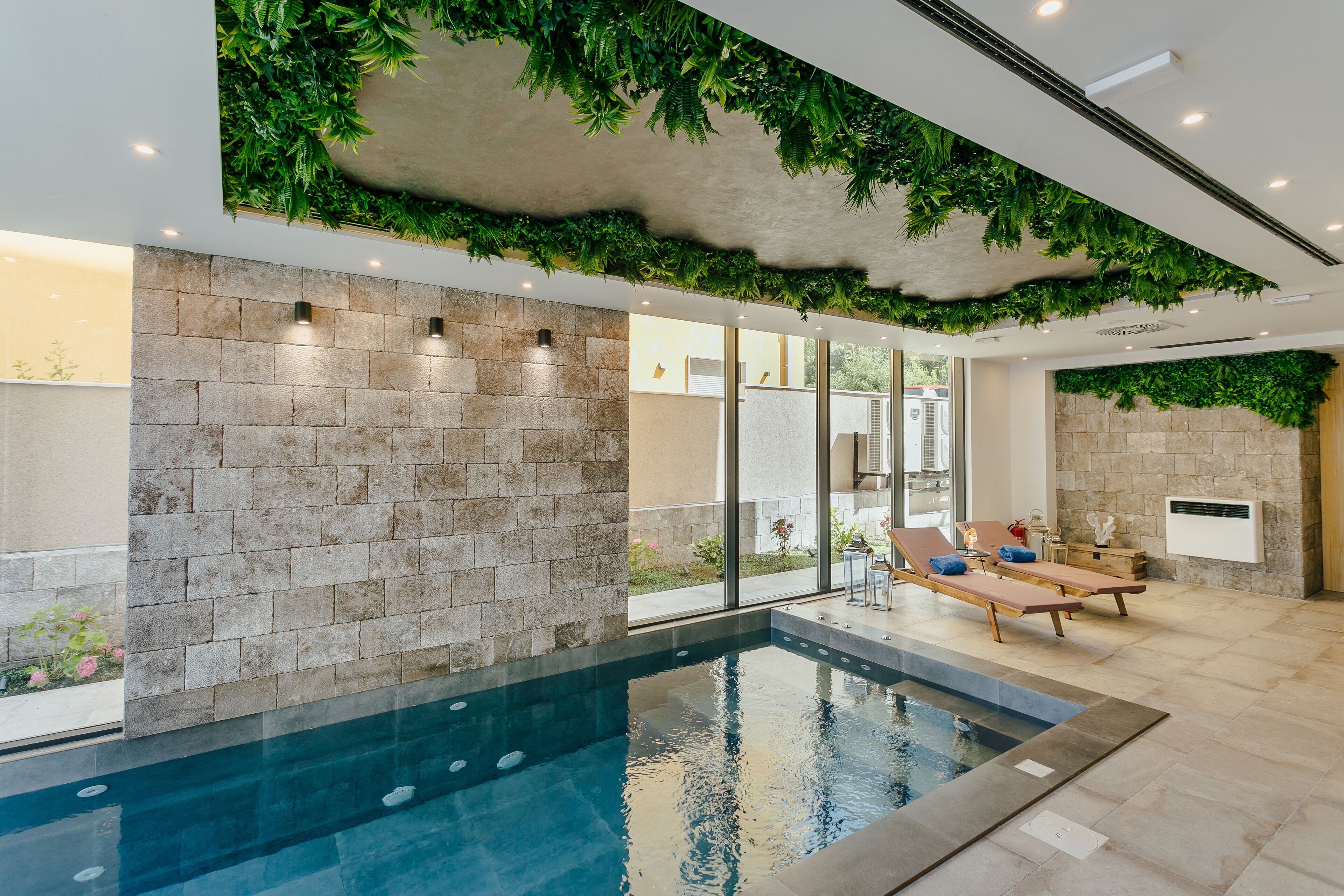 An indoor swimming pool with stone walls and ceiling greenery, lounge chairs, and a view of the outdoors.
