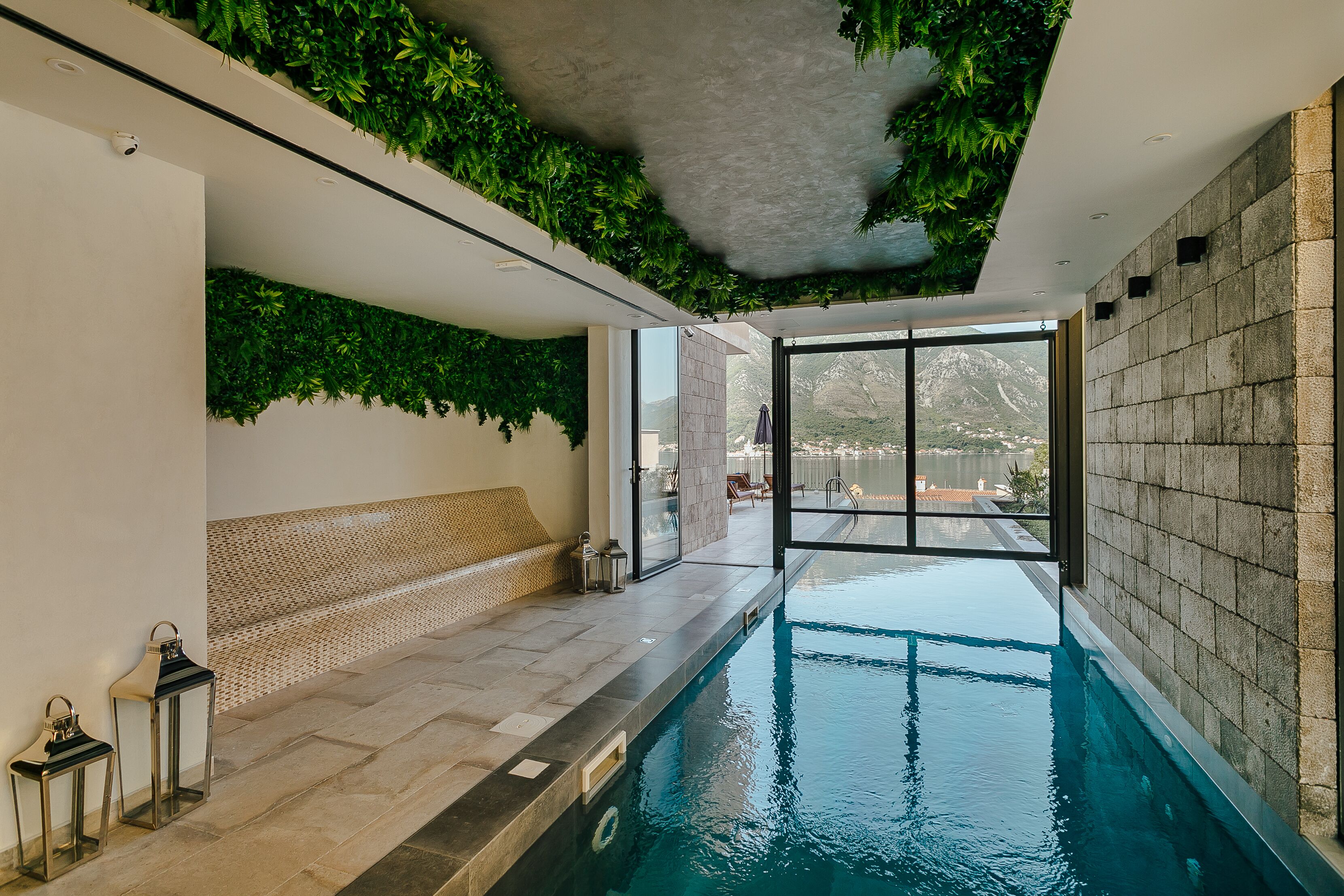 An indoor swimming pool with a view of mountains, a bench, and a ceiling with green foliage.