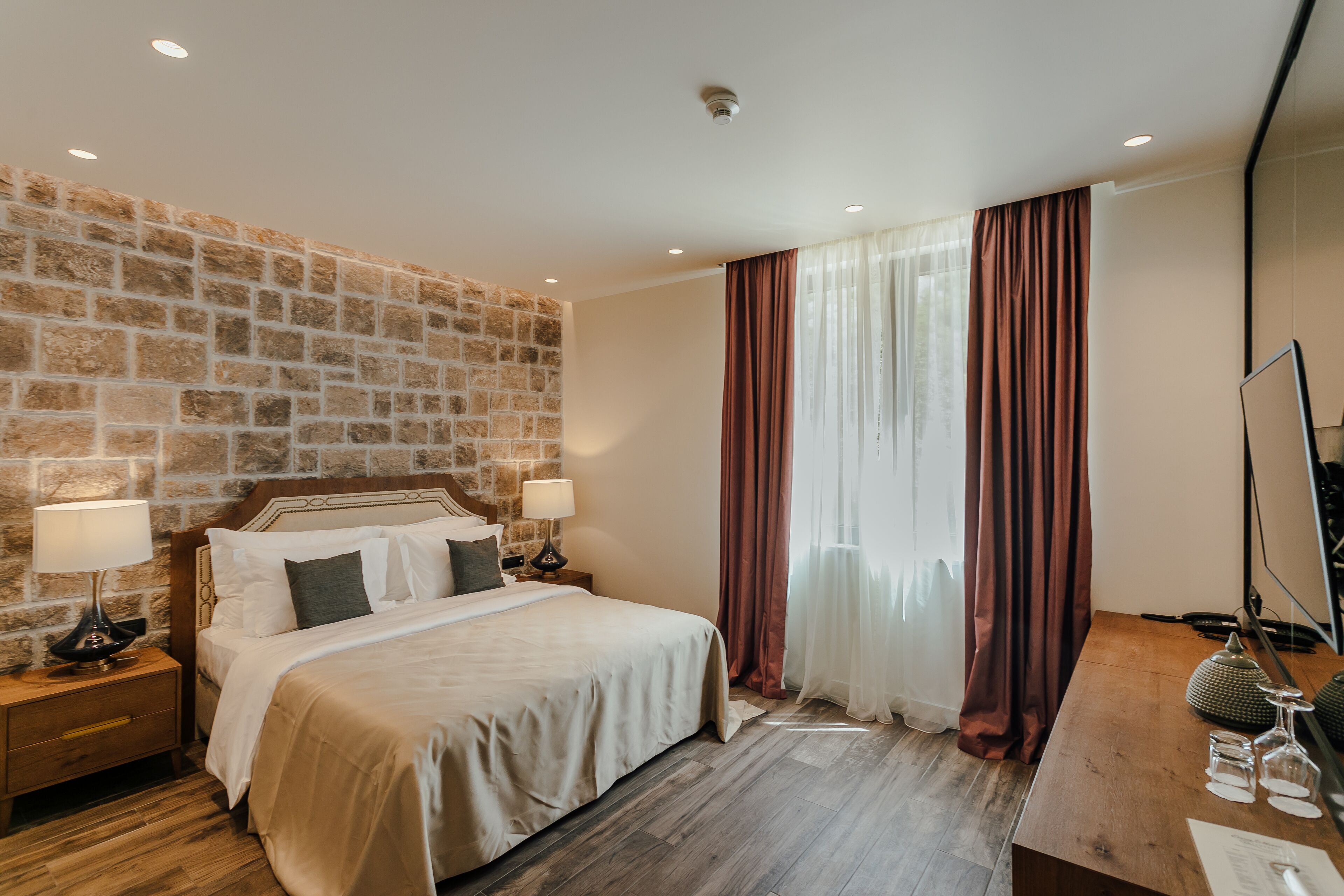 A hotel room with a stone accent wall, a large bed with white linens, two bedside lamps, a wooden nightstand, a desk with a mirror, and a window with sheer curtains.