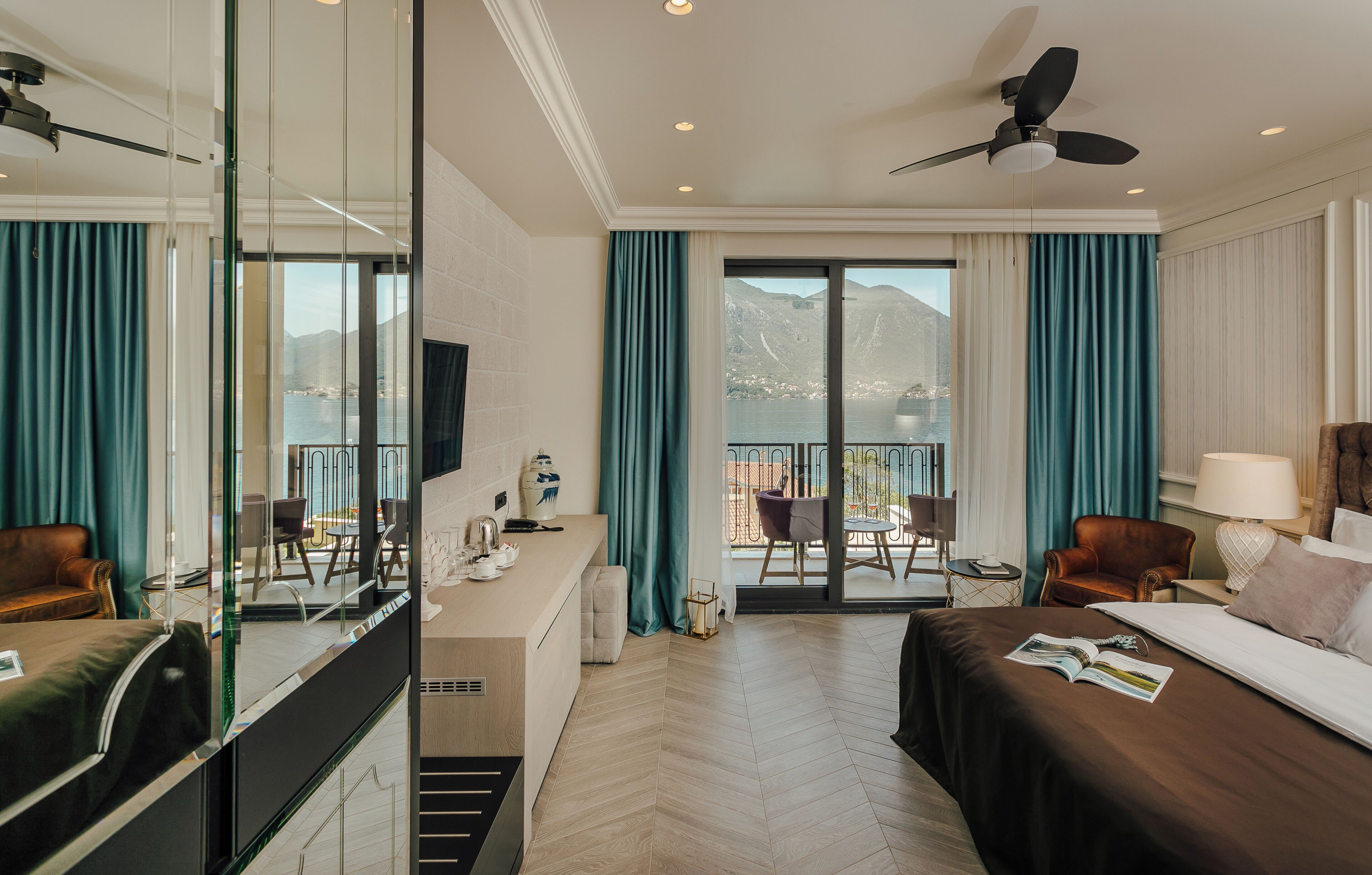A hotel room with a bed, a desk, a chair, a sofa, a balcony with a view of mountains, and a ceiling fan.