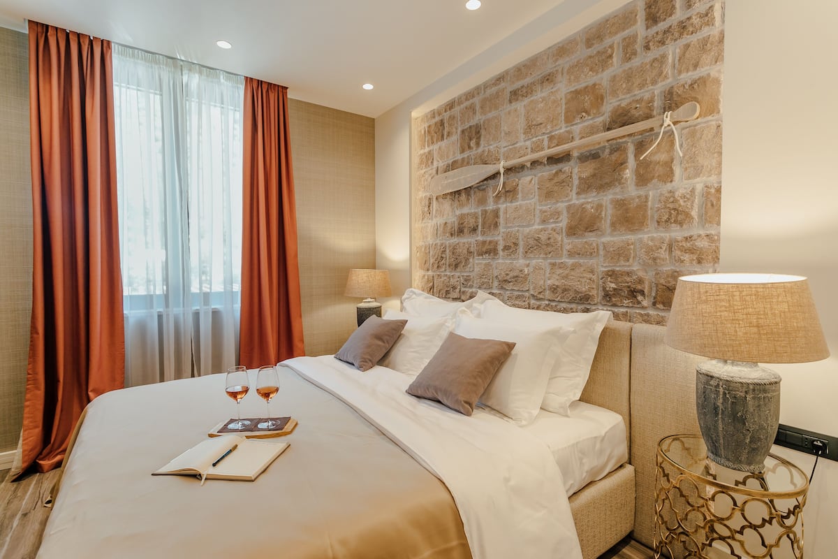 Una habitación de hotel con una cama grande, una pared de piedra, una mesita de madera con una lámpara y una ventana con cortinas.