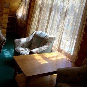 Interior acogedor de una cabaña con piso de madera, un sillón estampado con motivos florales, una pequeña mesa de madera y una ventana con cortinas de encaje.