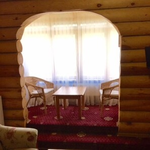 Una habitación acogedora con detalles de madera, que incluye una pequeña mesa y dos sillas, un área con alfombra roja y un amplio ventanal con cortinas traslúcidas.