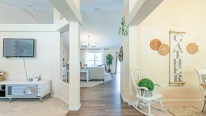Uma sala de estar ampla com piso de madeira, um sofá branco, uma mesa de jantar e uma TV fixada na parede.
