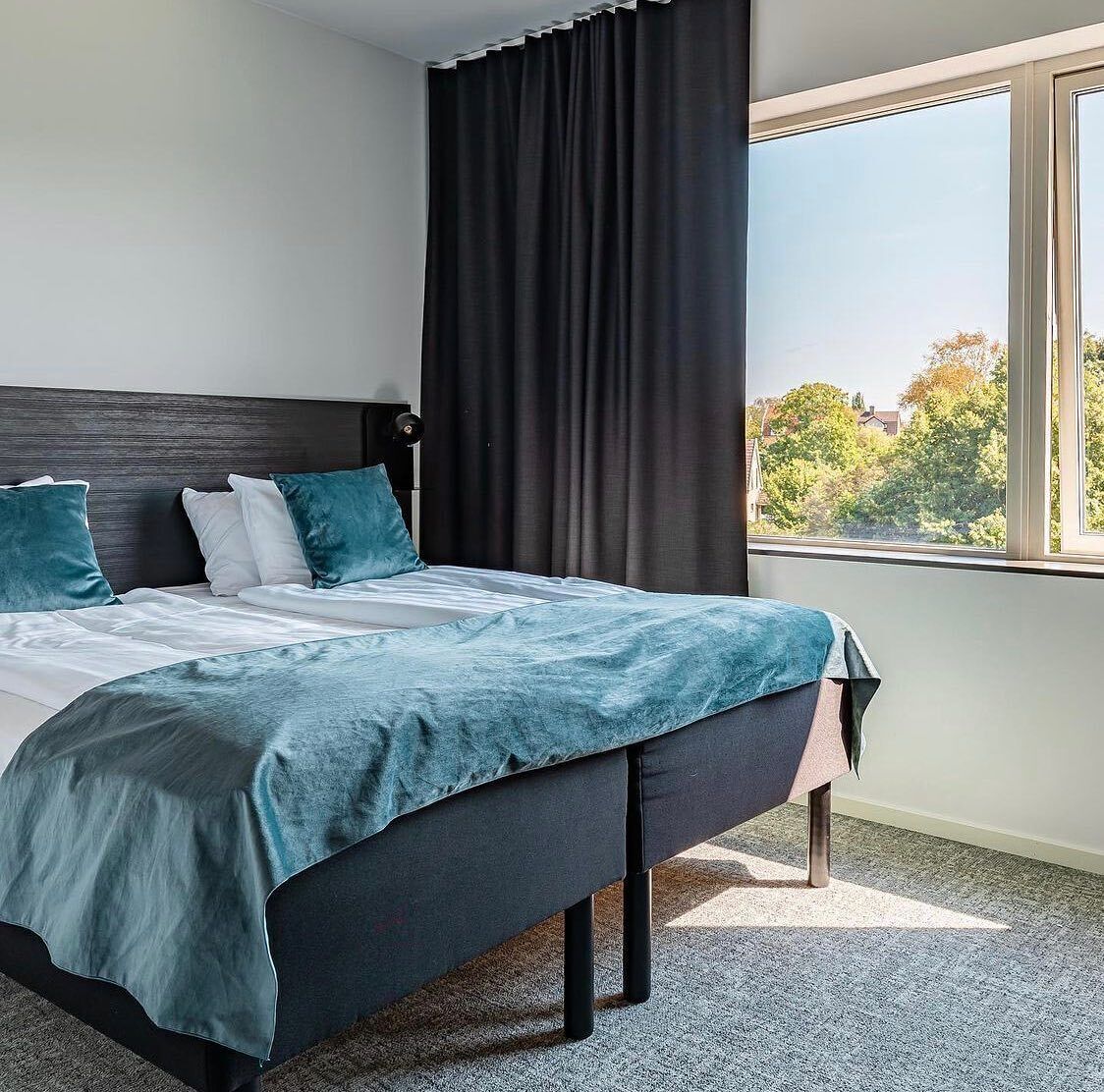 A modern hotel room with a large bed, dark curtains, and a view of trees through a window.