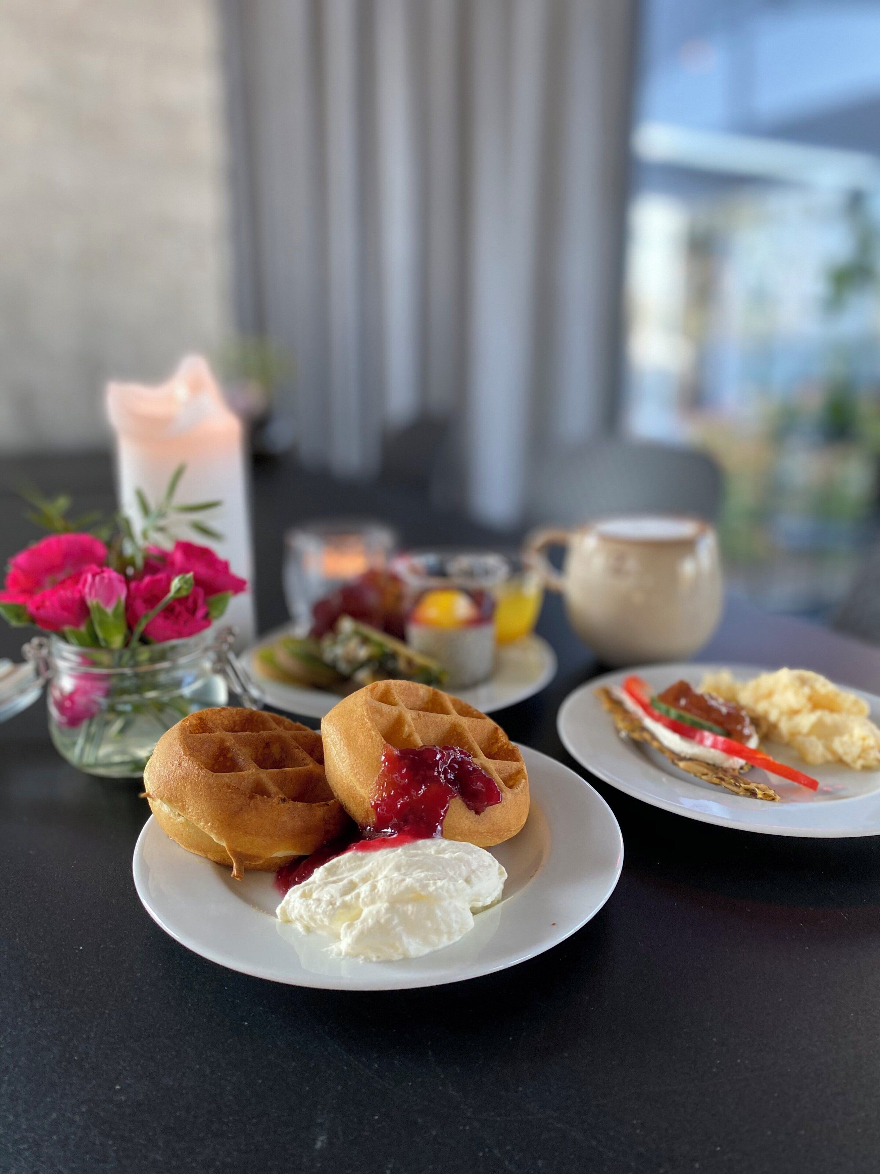 A breakfast setting with waffles, cream, and a variety of fruits on plates, a candle, and a vase of flowers.