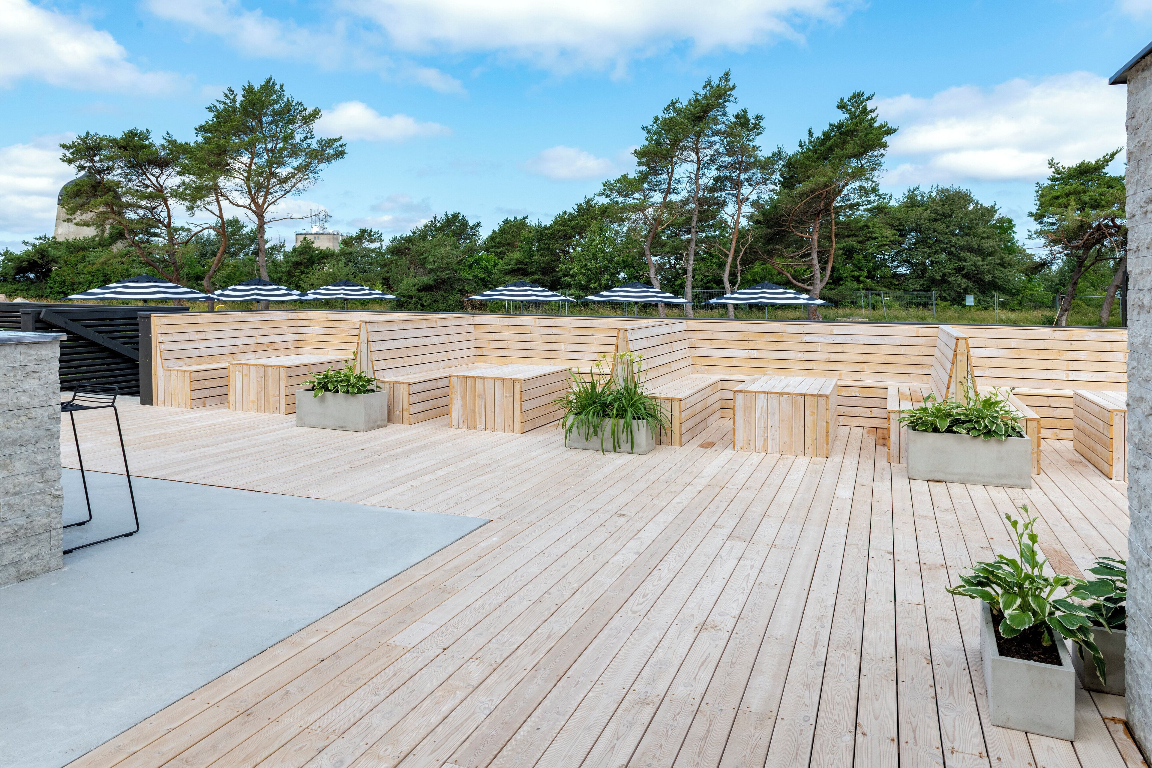Eine Holzterrasse mit Sitzbereichen und Pflanzkübeln, umgeben von Bäumen und blauem Himmel.