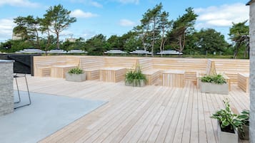 Eine Holzterrasse mit Sitzbereichen und Pflanzkübeln, umgeben von Bäumen und blauem Himmel.