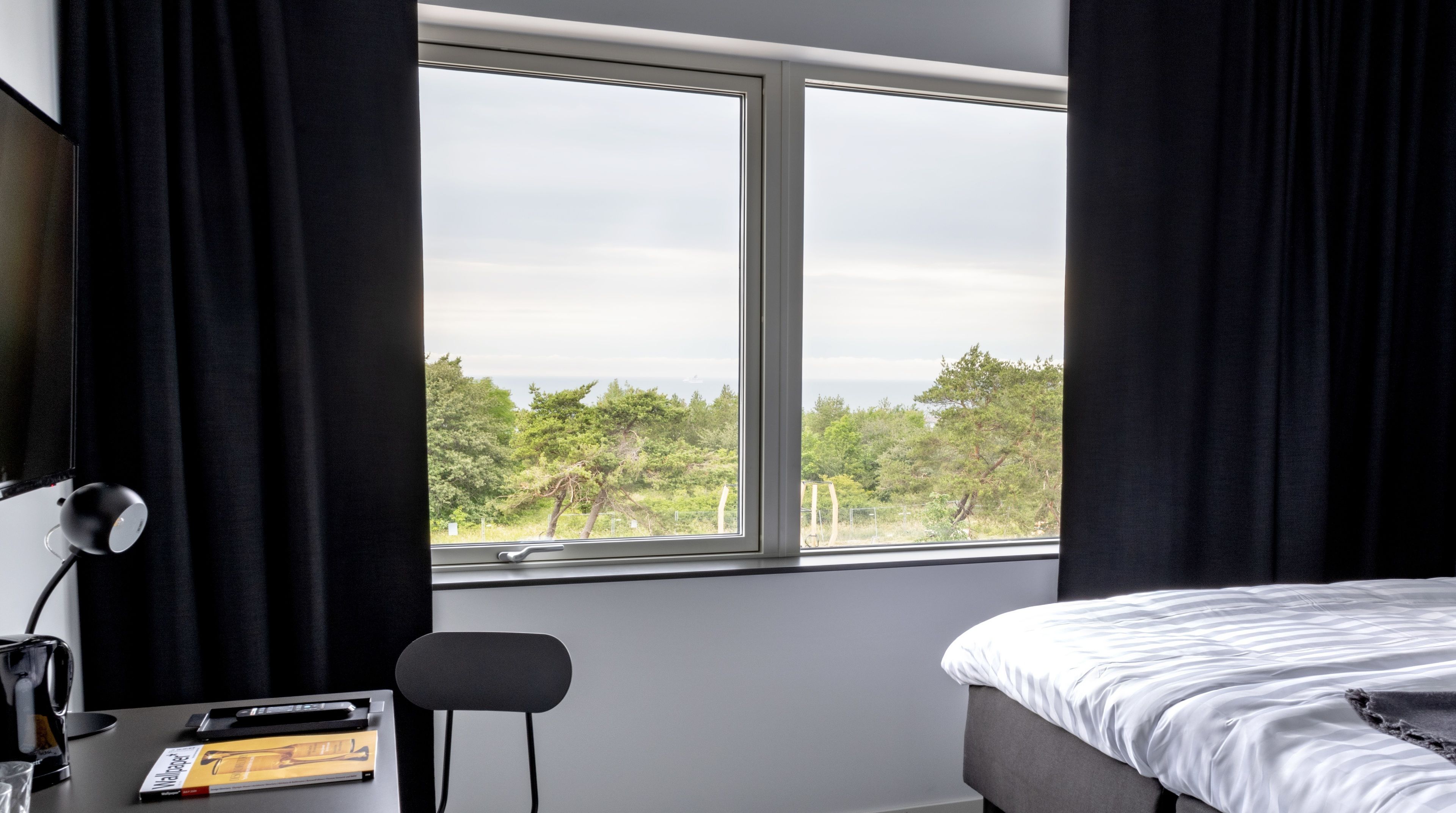 A bedroom with a large window, a bed, a desk with a lamp, and a chair.
