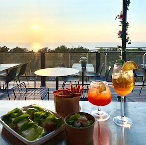 Una mesa dispuesta con ensalada, un bol de cóctel de camarones y un cóctel con un adorno de limón, con vista a un cuerpo de agua al atardecer.