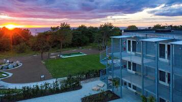 Un edificio moderno con fachada de vidrio, un jardín en la azotea y vistas al atardecer sobre un cuerpo de agua.