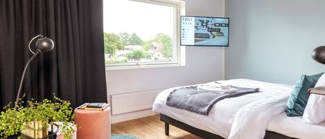 Un dormitorio con cama, ventana, planta, lámpara y pantalla mural.