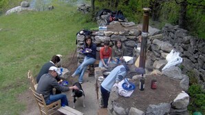 Un grupo de personas sentadas alrededor de un hogar de piedra con un fogón portátil, comiendo y relajándose al aire libre.