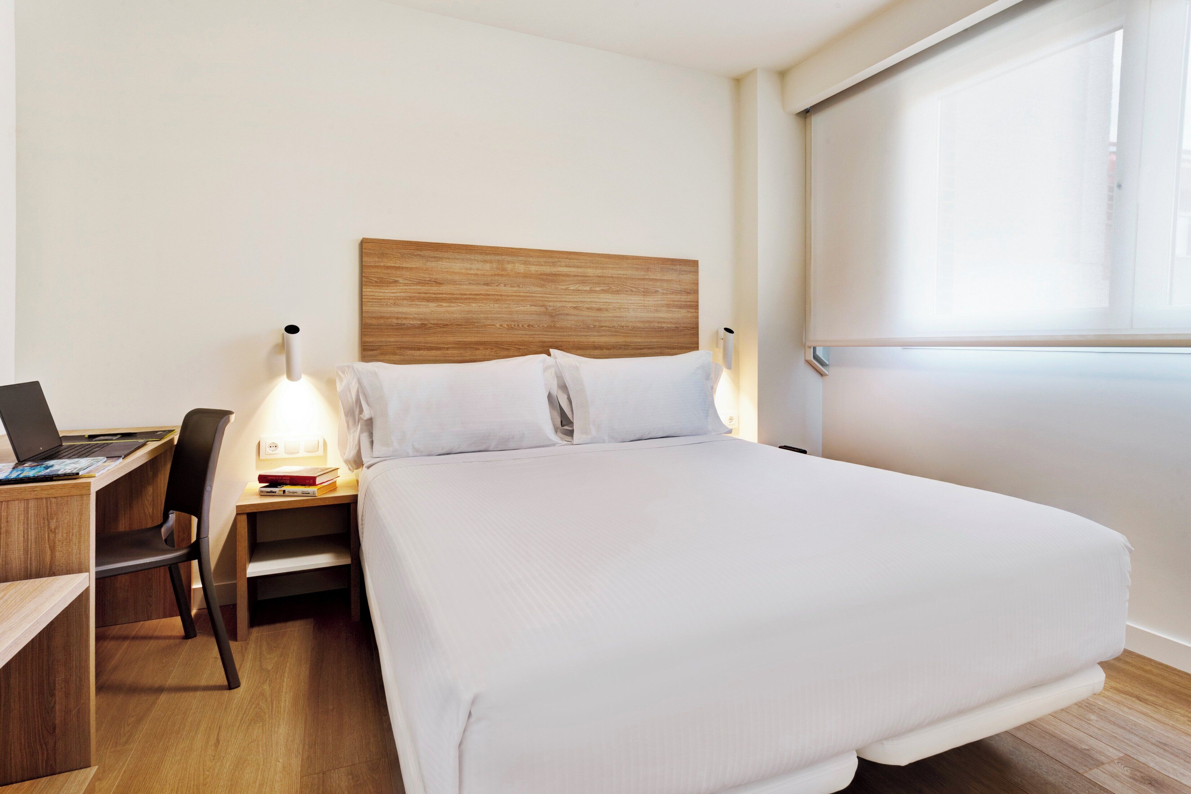 A hotel room with a large bed, a desk with a laptop, a chair, and a bedside lamp.