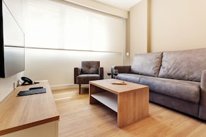 Una sala de estar moderna con una mesa de centro de madera, dos sofás grises, un televisor y un escritorio con teléfono y control remoto.