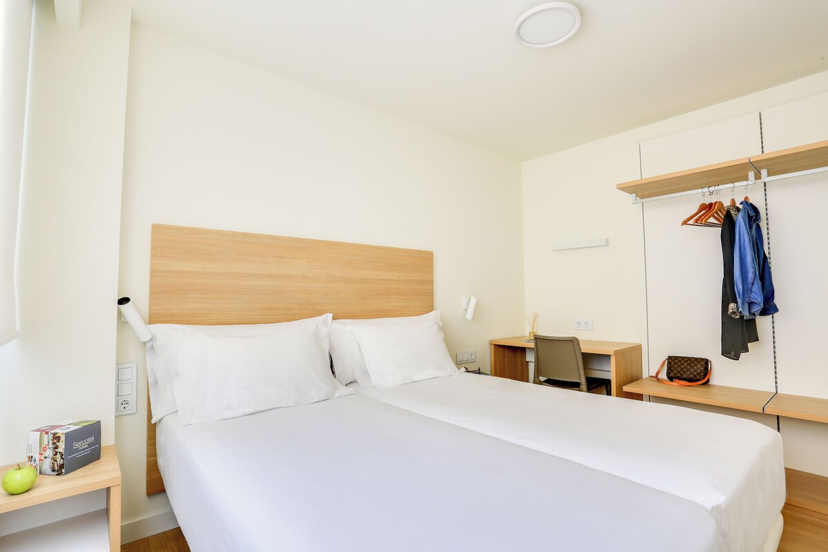 A hotel room with a bed, a desk, a chair, a hanging coat rack, and a small table with a bag and an apple.