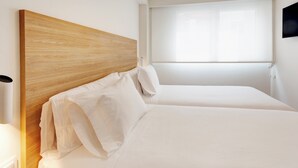 A hotel room with a wooden headboard, two beds, white linens, a bedside table, and a window with blinds.