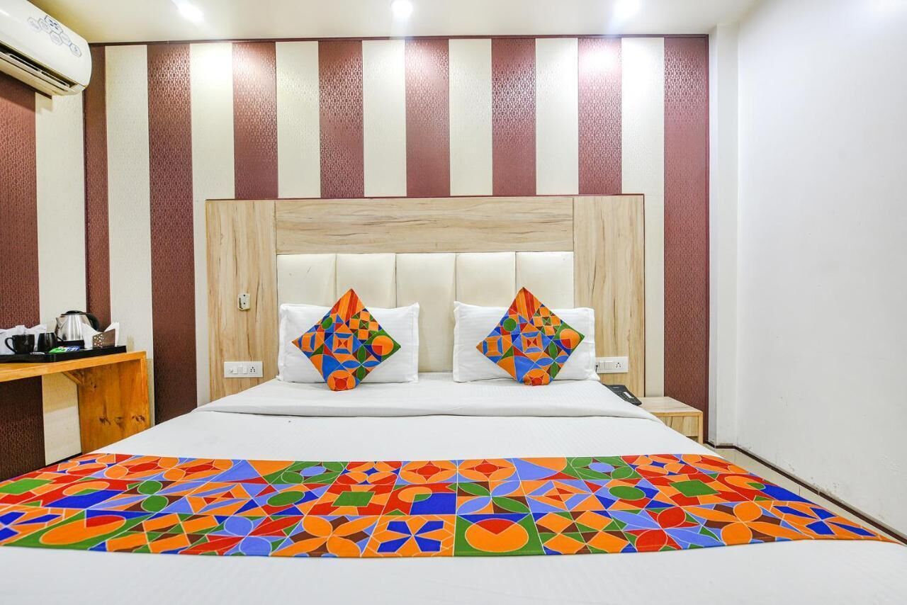 A hotel room with a striped wall, a wooden headboard, two pillows with colorful geometric patterns, a bed with a multicolored geometric-patterned bedspread, and a small wooden table with a kettle and cups.