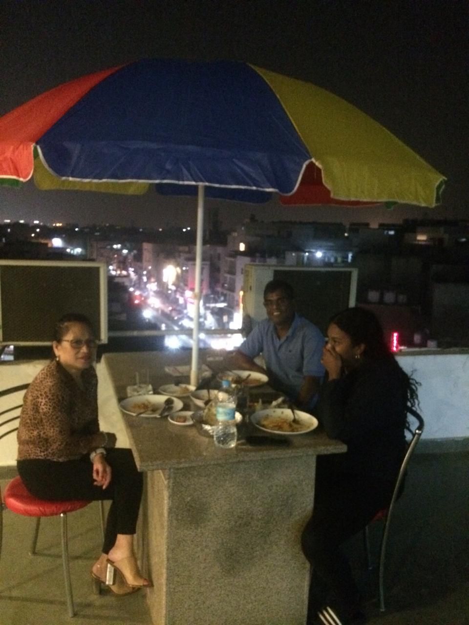 A group of people dining at an outdoor table with a colorful umbrella, surrounded by city lights.