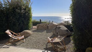 Une terrasse avec deux fauteuils de jardin en bois, de gros rochers et une vue sur la mer.