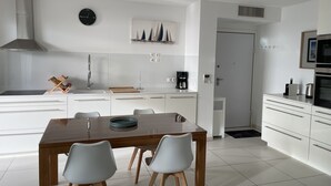 Une cuisine moderne avec des meubles blancs, une table à manger en bois accompagnée de quatre chaises, une machine à café et une horloge au mur.