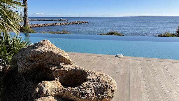 Un ciel d’un bleu limpide, une mer calme et une terrasse en bois avec un grand rocher au premier plan.