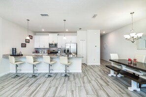Una cocina moderna con gabinetes blancos, una isla central con taburetes, electrodomésticos de acero inoxidable y una zona de comedor con una mesa de madera oscura y bancos.