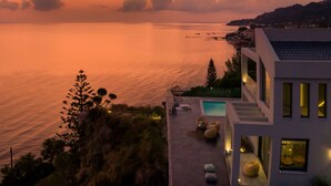 A modern building with a pool, outdoor seating, and a view of the sea at sunset. - Sueño LUXURY VILLA 180 Iconic Sea View (Zakynthos)