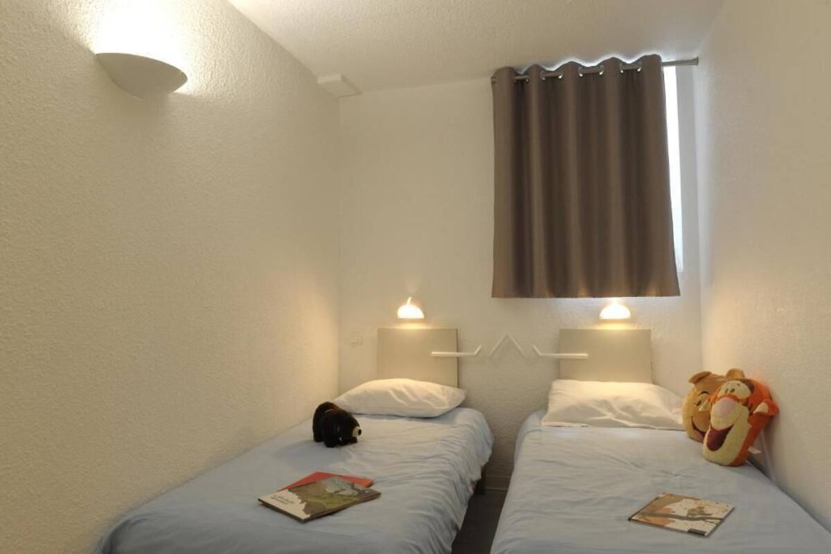 Una habitación pequeña y limpia con dos camas individuales, una ventana con cortinas y una lámpara fijada a la pared.