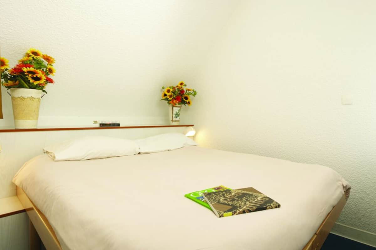 Habitación de hotel con cama, ropa de cama blanca, dos floreros y una revista sobre la cama.