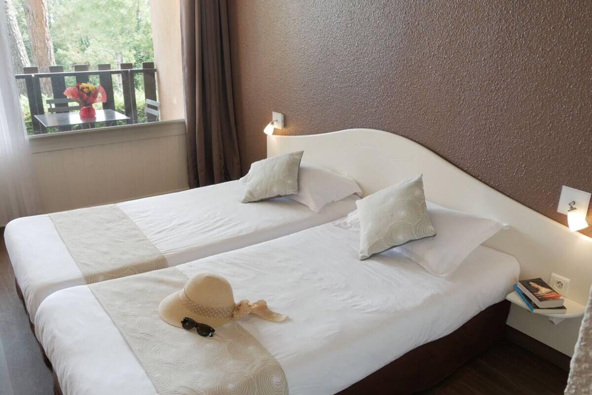 Cama doble con ropa blanca y dos almohadas, mesita de noche con lámpara, y ventana con vista a zonas verdes.