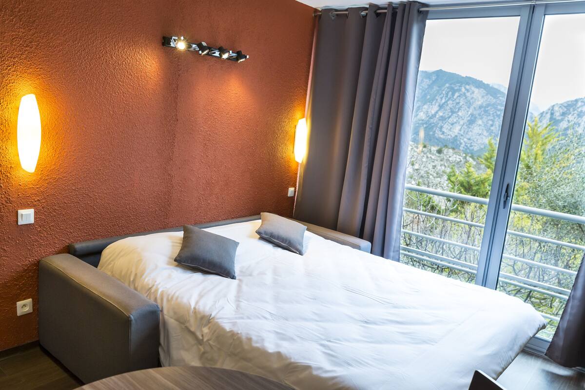 Una habitación de hotel con una cama, dos apliques, una ventana con cortinas y vistas a las montañas.