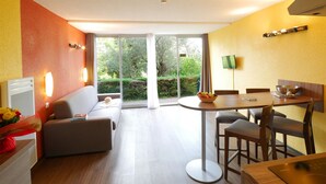 A modern hotel room with a sofa, dining table, and chairs, a small kitchen area, and a sliding glass door leading to a garden.