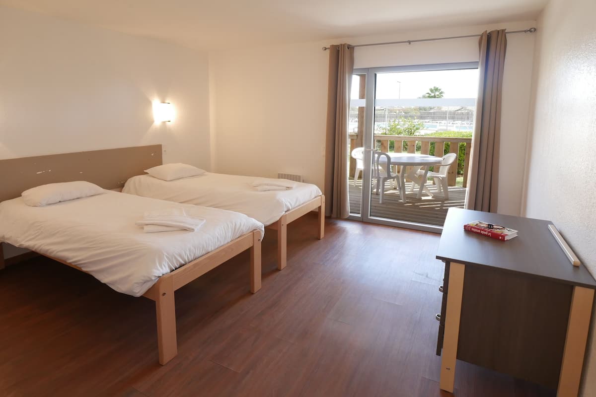 Une chambre d’hôtel avec deux lits simples, un petit bureau et une porte-fenêtre coulissante donnant sur un espace extérieur aménagé avec une table et des chaises.