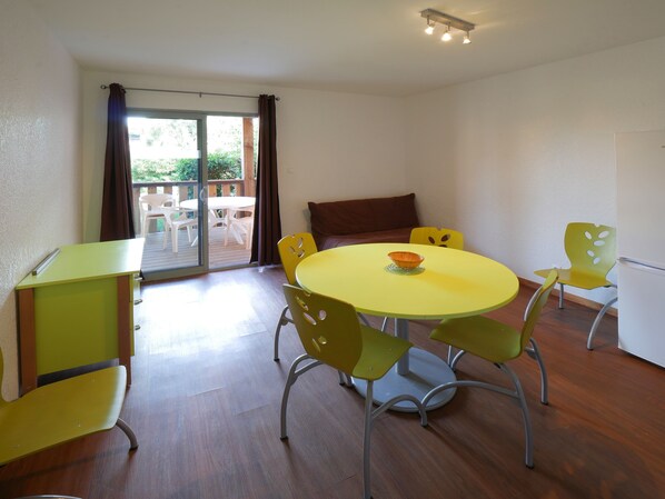 Una habitación con una mesa amarilla y sillas verdes, un ventanal corredero a un área exterior y un refrigerador blanco.