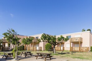 Una hilera de edificios modernos de dos pisos con balcones, mesas de picnic y árboles en un entorno exterior soleado.