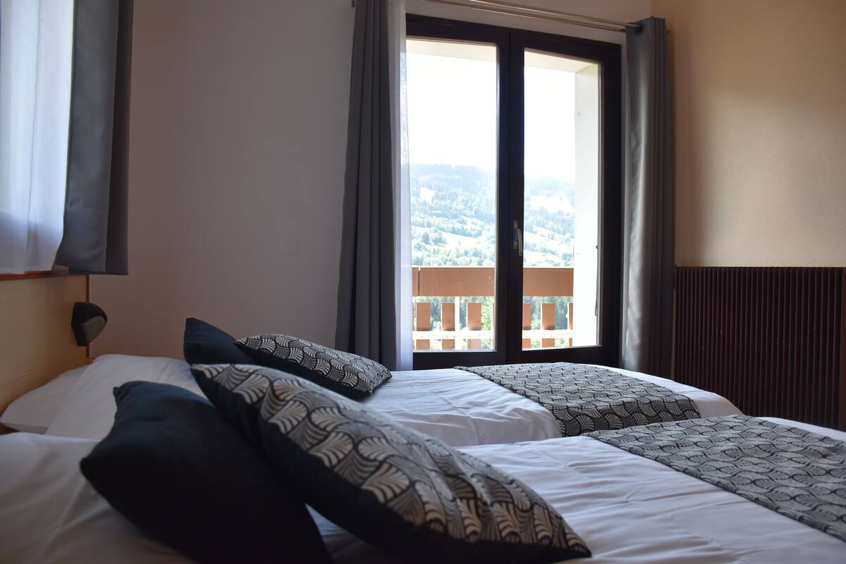 A hotel room with two beds, a window with a view of a balcony, and a wall-mounted lamp.