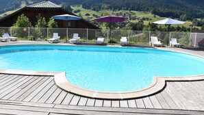 A swimming pool with lounge chairs and umbrellas, surrounded by a wooden deck and a metal fence, with a mountain view in the background. - Les Essertets - Vacances ULVF (Praz-sur-Arly)