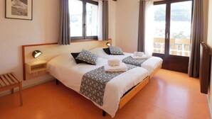 A double bed with patterned pillows, a bedside table with a lamp, and a window with curtains. - Les Essertets - Vacances ULVF (Praz-sur-Arly)